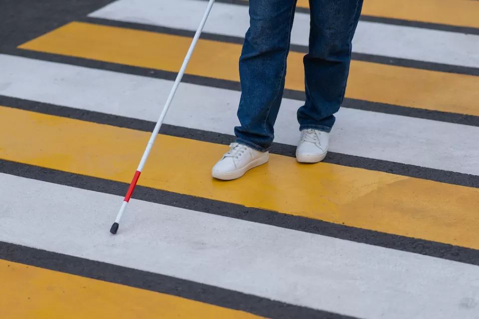 Close-up of person with vision impairment wearing jeans and white sneakers crossing yellow and white striped crosswalk with cane.